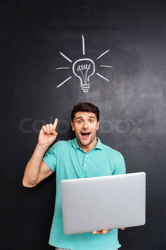 Excited young man pointing finger up and holding laptop over blackboard background with drawn light bulb, stock photo