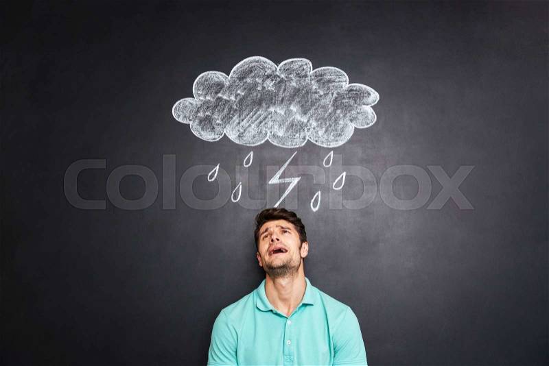 Desperate depressed young man standing and crying under raincloud with lightning and rain drawn on chalkboard background, stock photo