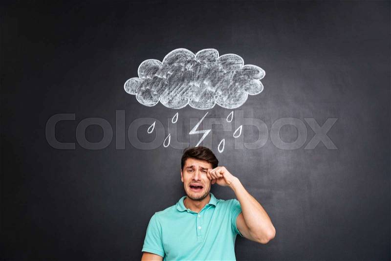 Sad young man crying over black blackboard with drawn raincloud, stock photo
