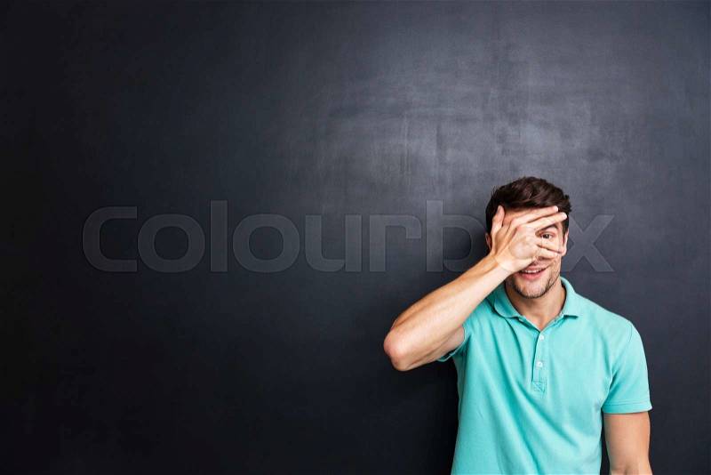 Casual smiling man covering his eyes with fingers isolated over black chalkboard, stock photo