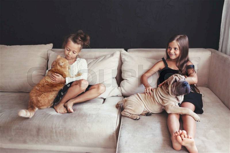 Home portrait of two cute children hugging with ginger cat and puppy of Chinese Shar Pei dog on the sofa against black wall, stock photo