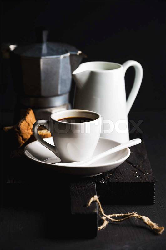 Italian coffee set for breakfast. Cup of hot espresso, creamer with milk, cantucci and moka pot on dark rustic wooden board over black background, selective focus, vertical composition, stock photo