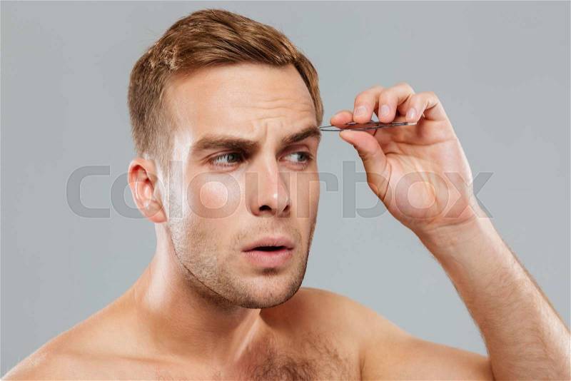 Closeup of concentrated young man removing eyebrow hairs with tweezers, stock photo