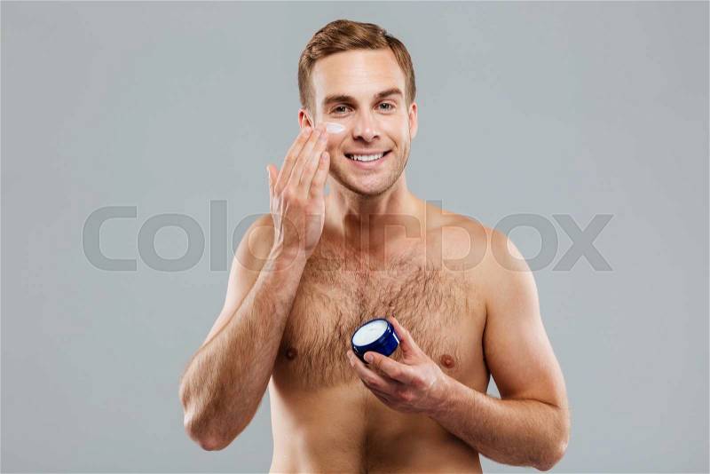 Young handsome man putting on cream lotion on face isolated on the gray background, stock photo
