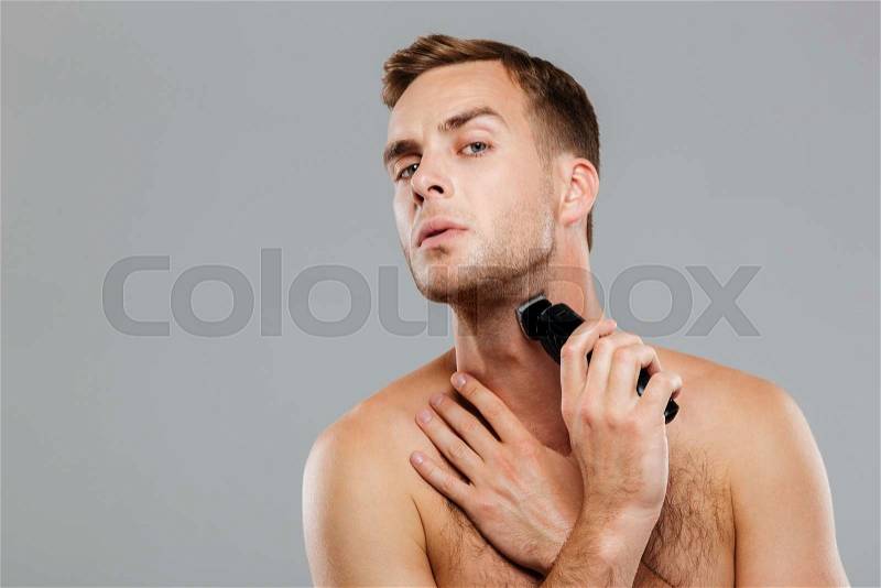 Young happy man shaving with electric razor isolated on the gray background, stock photo