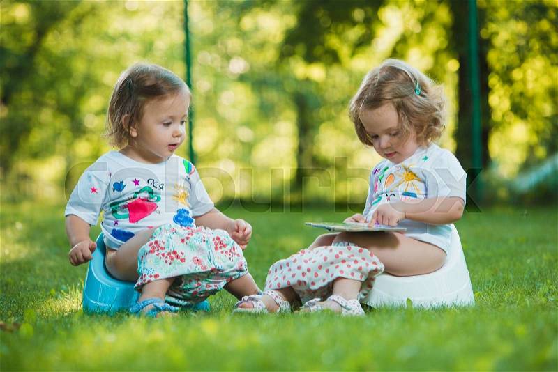 The two little baby girls two-year old sitting on pottys on green grass of lawn, stock photo
