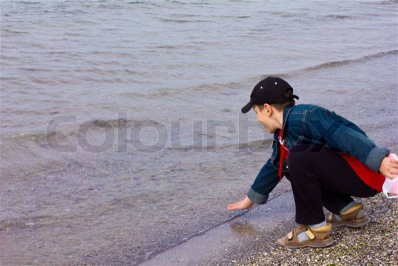 10 year old boy in denim jacket, cap, sandals squatting on the beach and the water touches his hand, stock photo
