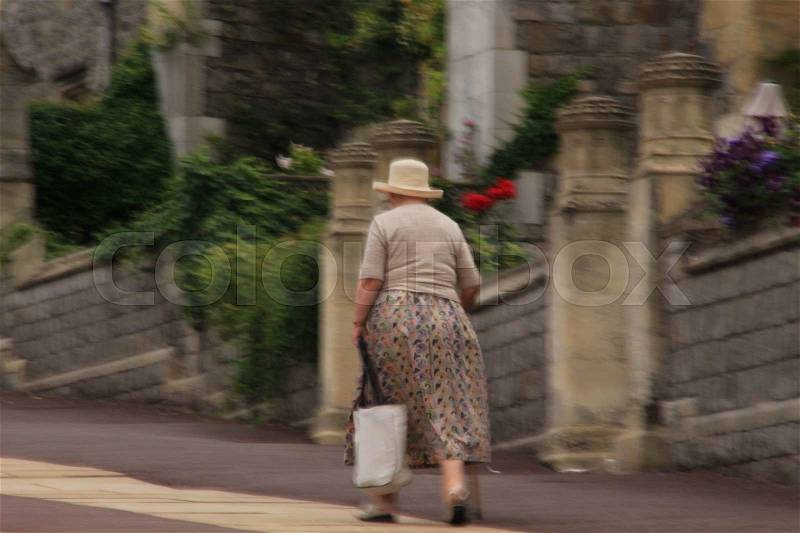 The dignified lady with hat, walking stick and shopping bag is walking on the street in Windsor Castle in England in the summer, stock photo