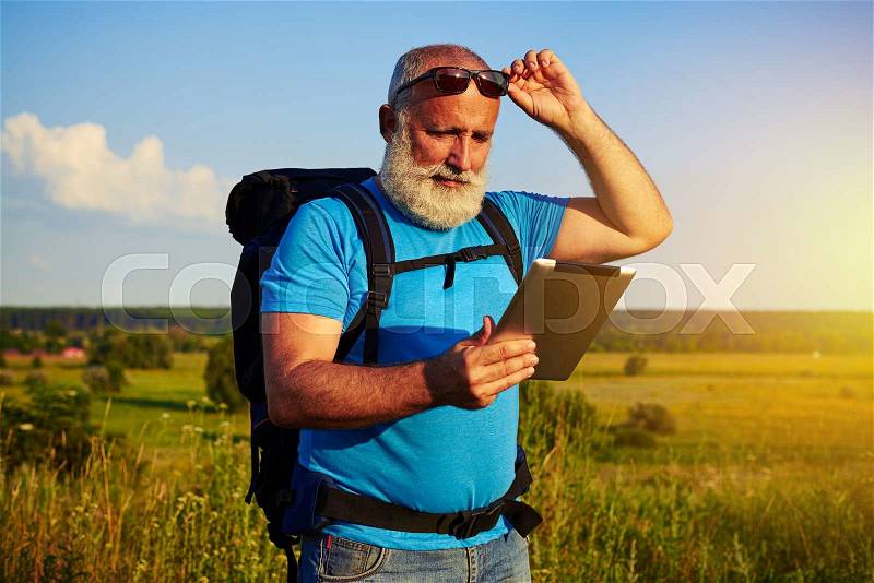 Handsome man with white beard in sunglasses with rucksack is using data tablet against sunlit field background, stock photo