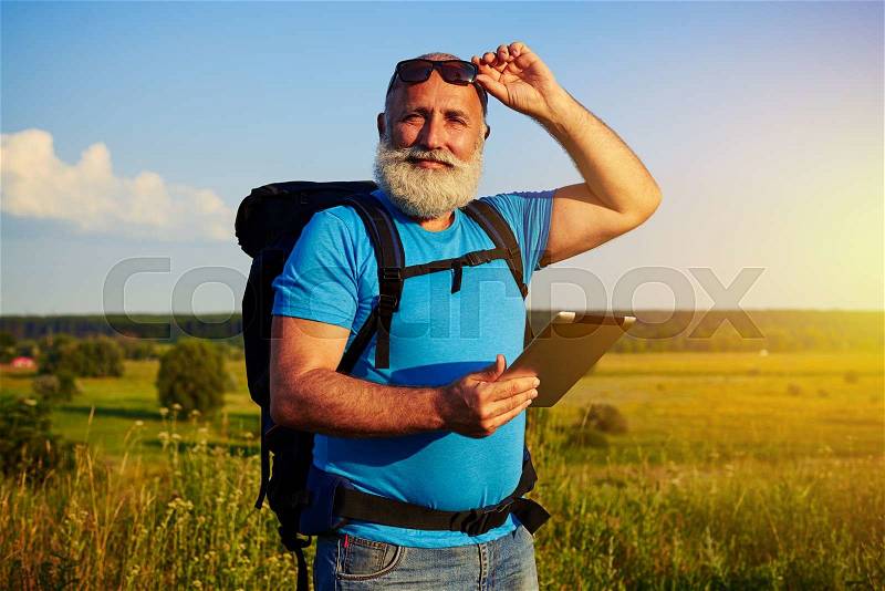 Portrait of handsome man with white beard carrying a rucksack and holding a data tablet against sunset field background, stock photo