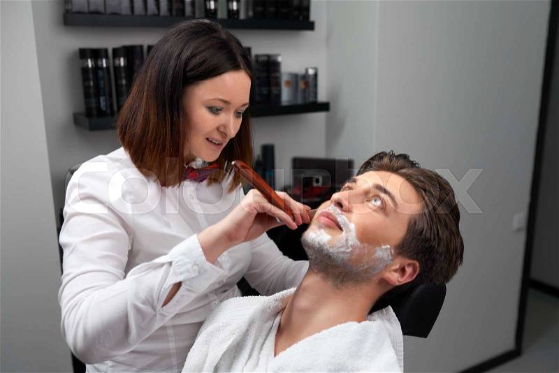 The barber woman shaves client\'s beard in the barber shop, stock photo