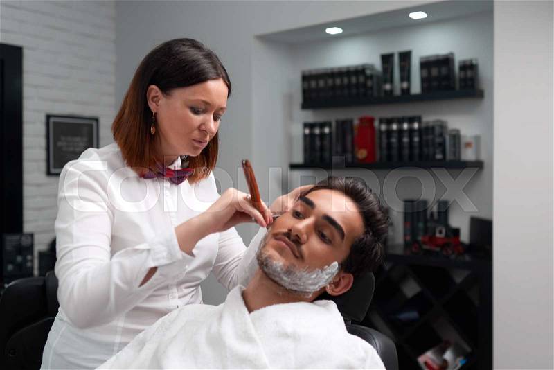 The barber woman carefully shaves client\'s beard in the barber shop, stock photo