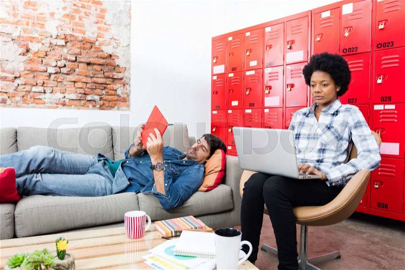 Two colleagues man lying on coach using tablet woman working with laptop modern coworking office business people casual wear, stock photo