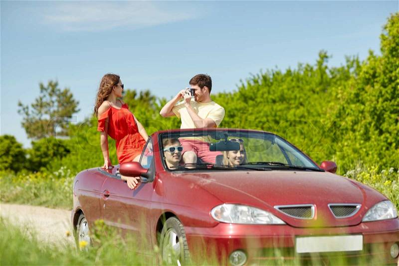Leisure, road trip, travel, summer holidays and people concept - happy friends driving in cabriolet car and taking picture by film camera, stock photo