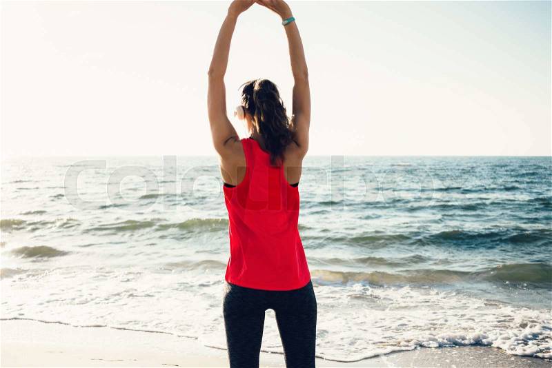 Girl in sportswear doing morning exercises on the beach and listening to music on headphones, view from the back, stock photo