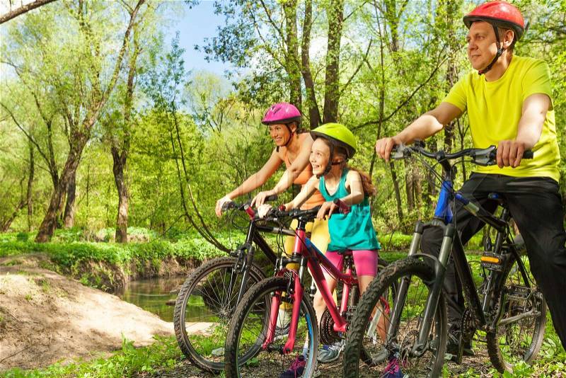 Happy cyclists, father, mother and girl, admiring the landscape of sunny spring wood riding their bikes, stock photo