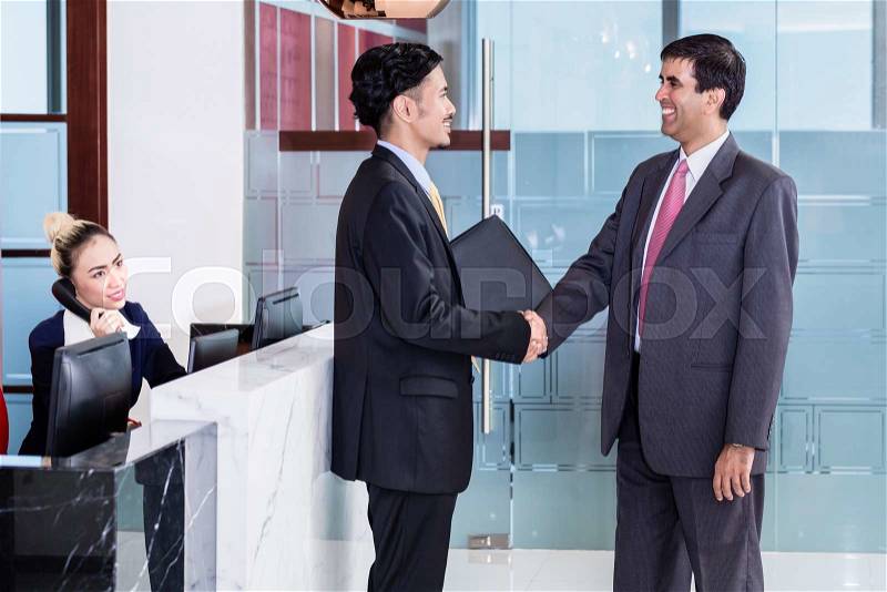 Indian business executive welcoming Asian associate in office lounge shaking hands, stock photo
