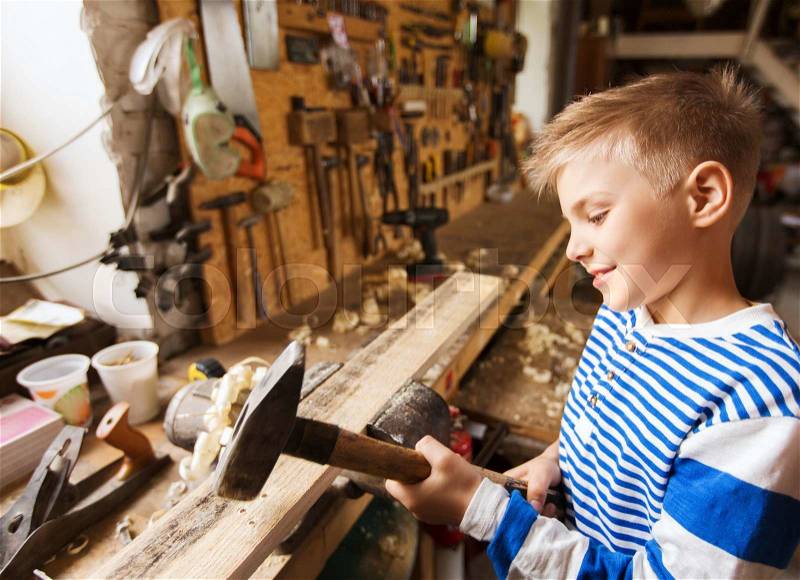 Carpentry, woodwork, building and people concept - happy little boy with hammer hammering wood plank at workshop, stock photo