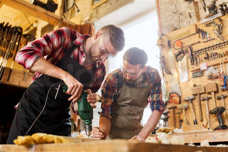 Profession, carpentry, woodwork and people concept - two carpenters with electric drill drilling wood plank at workshop, stock photo