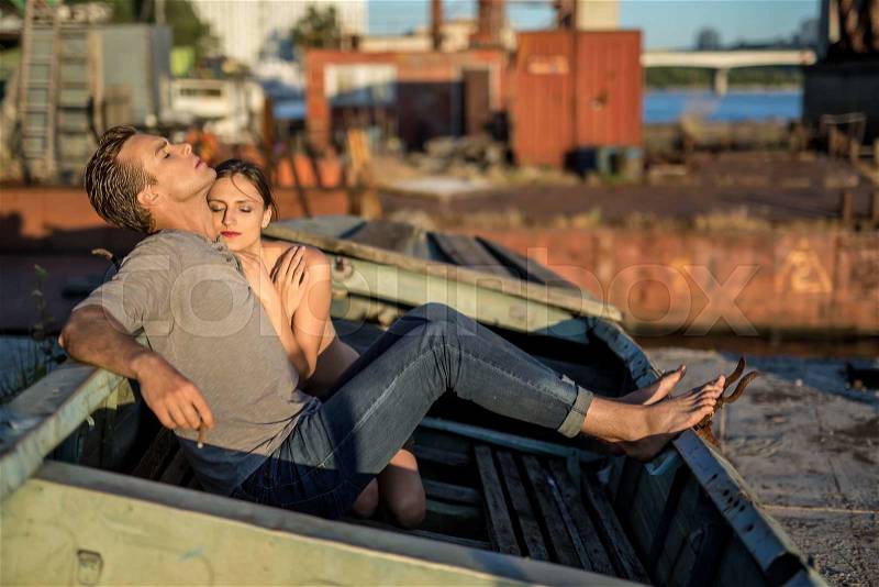 Attractive couple with closed eyes in the old boat outdoors in the industrial zone. Barefoot guy in a gray T-shirt and blue jeans smokes a cigarette. Nude girl cuddled up to him. Horizontal, stock photo