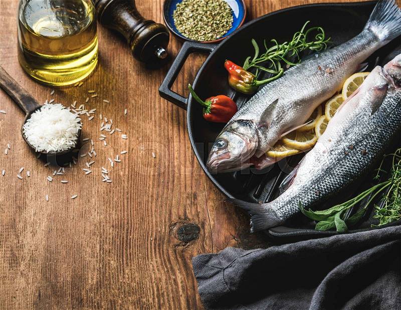 Ingredients for cookig healthy fish dinner. Raw uncooked seabass fish with rice, olive oil, lemon slices, herbs and spices on black grilling iron pan over rustic wooden background, top view, selective focus, copy space, horizontal composition, stock photo