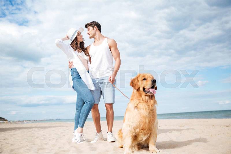 Dog sitting in front of happy young couple standing and hugging on the beach, stock photo