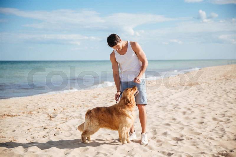 Handsome young man standing and playing with his dog on the beach, stock photo