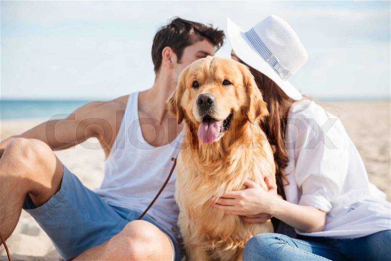 Beautiful young couple sitting and hiding behind the dog on the beach, stock photo