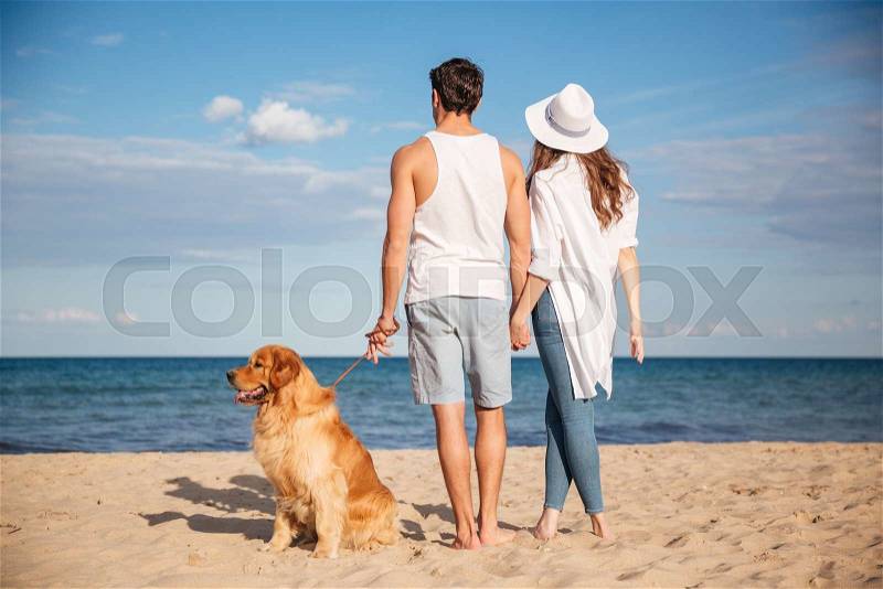 Back view of happy young couple walking with dog on the beach, stock photo