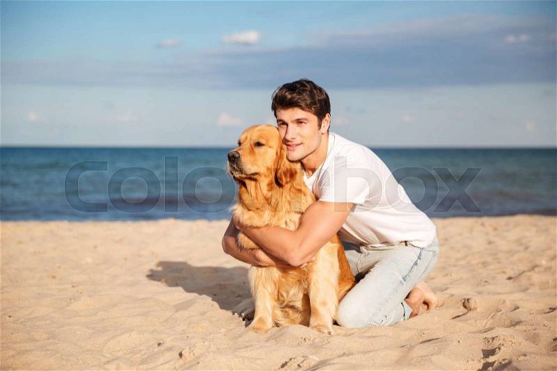 Handsome young man sitting and hugging his dog on the beach, stock photo