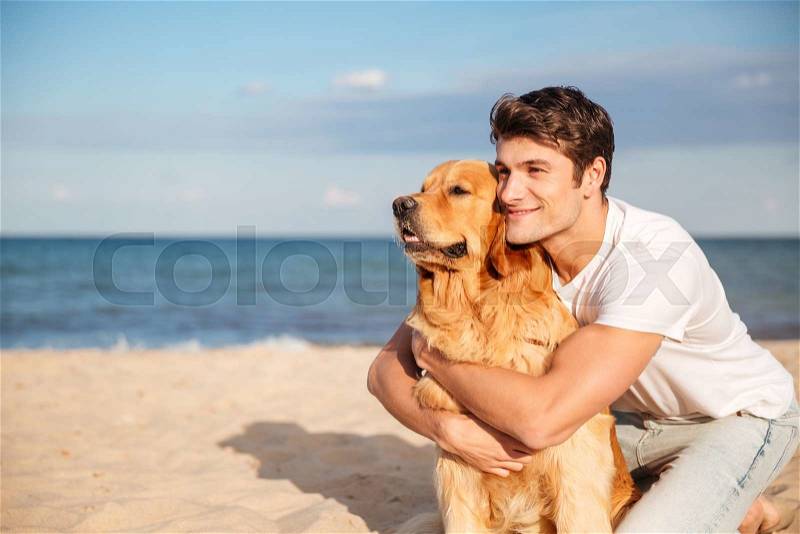 Smiling attractive young man hugging his dog on the beach in summer, stock photo