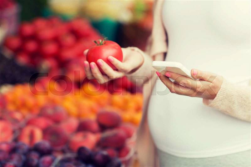 Sale, shopping, food, pregnancy and people concept - close up of pregnant woman with smartphone and tomato choosing vegetables at street market, stock photo