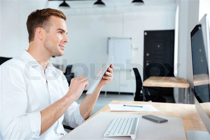 Smiling young businessman using tablet and computer at workplace, stock photo