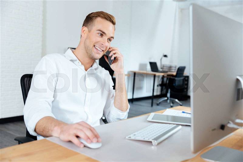 Smiling young businessman working with computer and talking on cell phone at workplace, stock photo