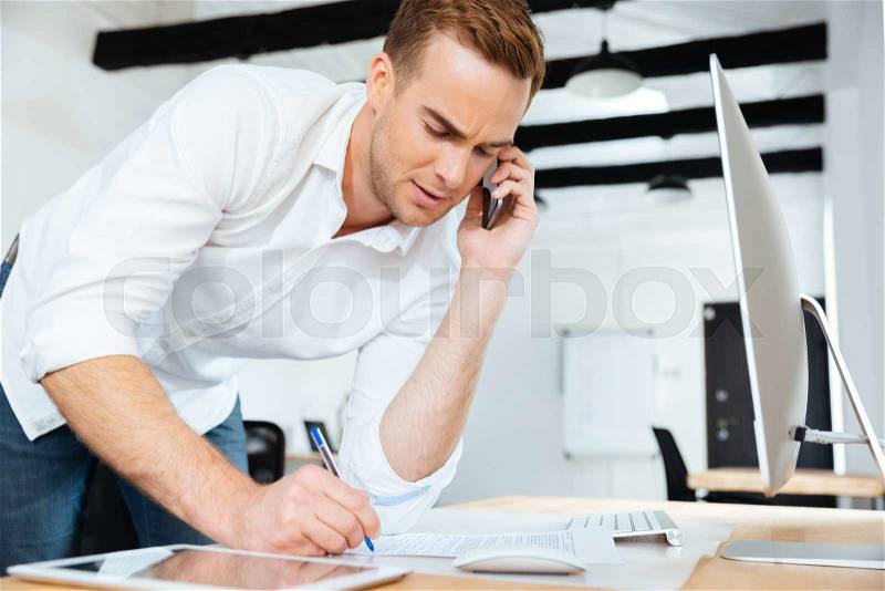 Handsome young businessman talking on cell phone and writing in office, stock photo