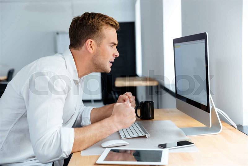 Mad irritated young businessman sitting and working with blank screen computer in office, stock photo