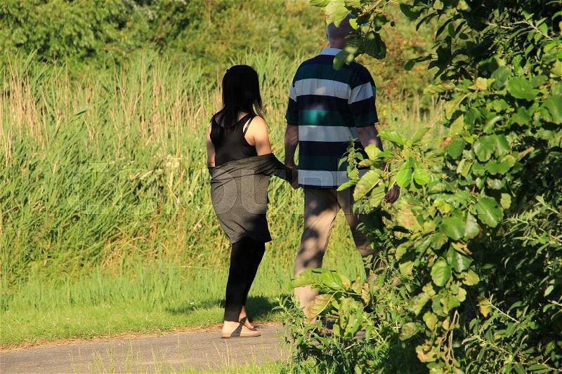 Couple, the man behind the leaves of the tree and the woman are walking hand in hand in the park at the country side in the summer, stock photo