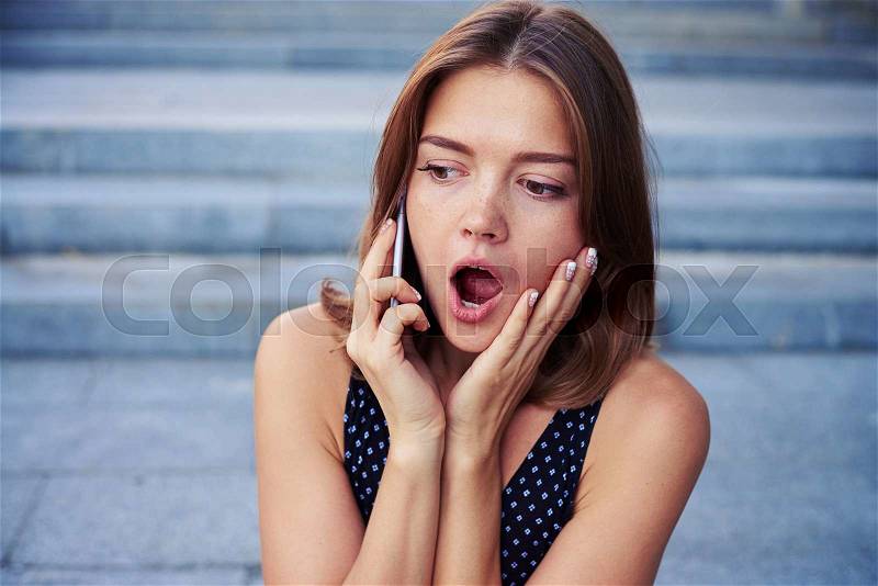 Beautiful woman opened her mouth in surprise while talking on the mobile phone on the street stairs, stock photo
