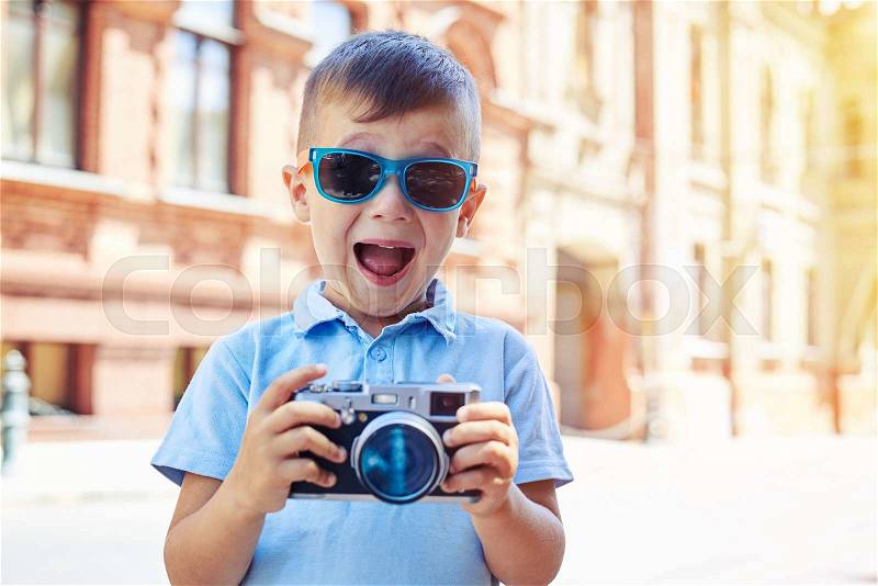 Small boy in sunglasses is making excited face while holding a photo camera and trying to take a shot during walk in historical part of the city, stock photo