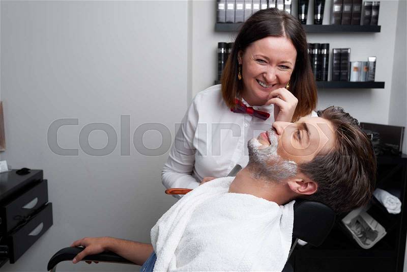 The laughing barber woman shaves client\'s beard and chin in the barber shop, stock photo