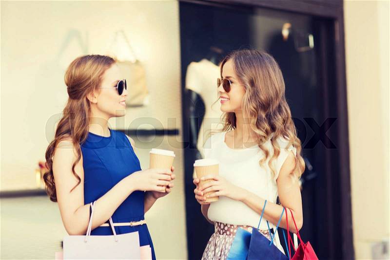 Sale, consumerism and people concept - happy young women with shopping bags and coffee paper cups talking at shop window in city, stock photo