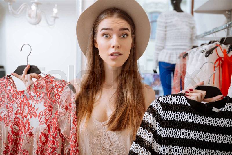 Confused pretty young woman choosing dress in clothing store, stock photo