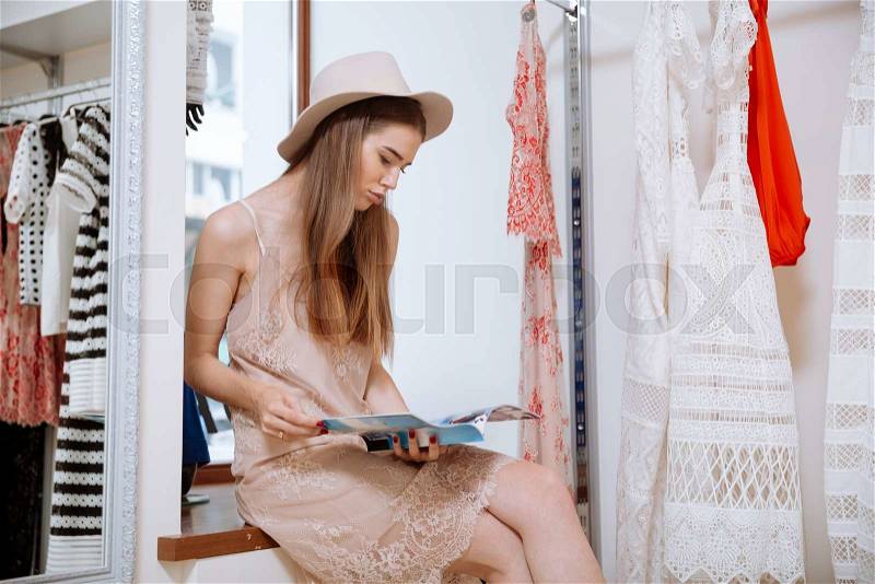 Relaxed attractive young woman in hat sitting and reading magazine in clothing store, stock photo