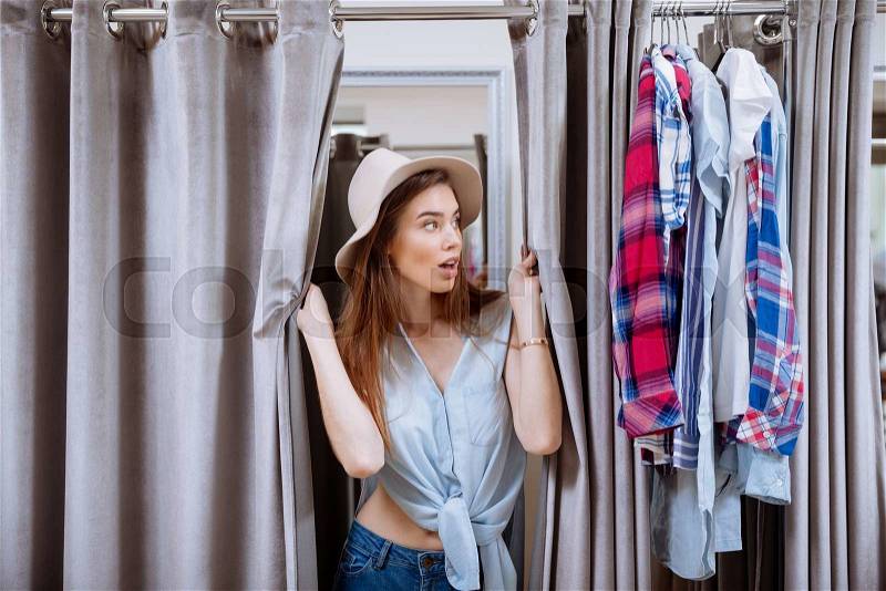 Surprised young woman trying on clothes and looking out of dressing room, stock photo