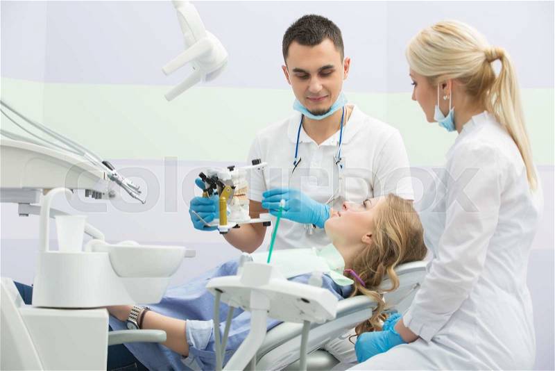 Nice girl in blue shirt and patient bib on the patient chair in the dental cabinet. Near her sits a male dentist and a female assistant. They both wear white uniform with blue latex gloves and blue masks. Dentist also has binocular loupes. He holds an art