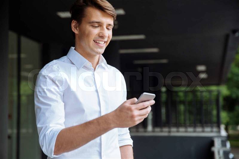 Happy attractive young businessman using smartphone and smiling near business center, stock photo
