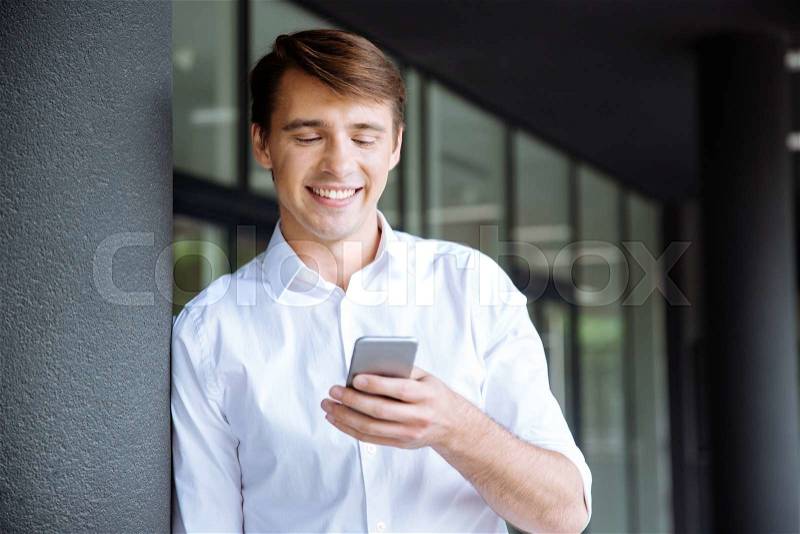 Smiling young businessman standing and using cell phone, stock photo