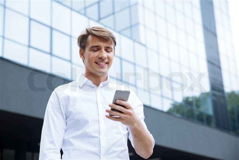 Smiling young businessman using mobile phone near business center, stock photo