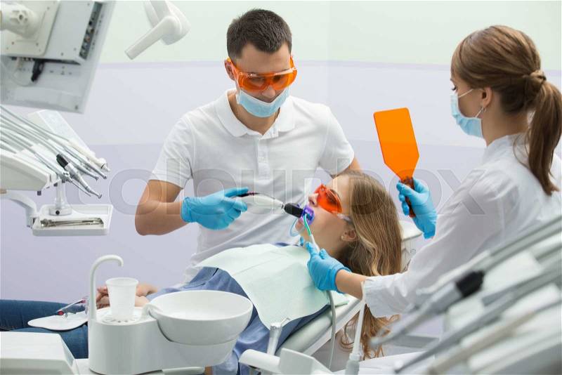 Nice girl in blue shirt, jeans and patient bib on the patient chair in the dental cabinet. Next to her there is a male dentist and a female assistant. They both wear white uniform with blue latex gloves and blue masks. Patient and dentist wear UV protecti