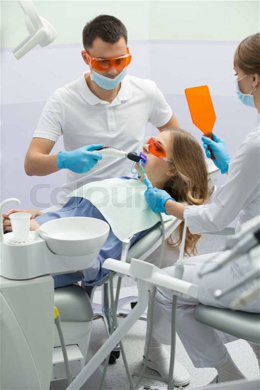 Cute girl in blue shirt and patient bib on the patient chair in the dental cabinet. Next to her there is a male dentist and a female assistant. They both wear white uniform with blue latex gloves and blue masks. Patient and dentist wear UV protective eyew